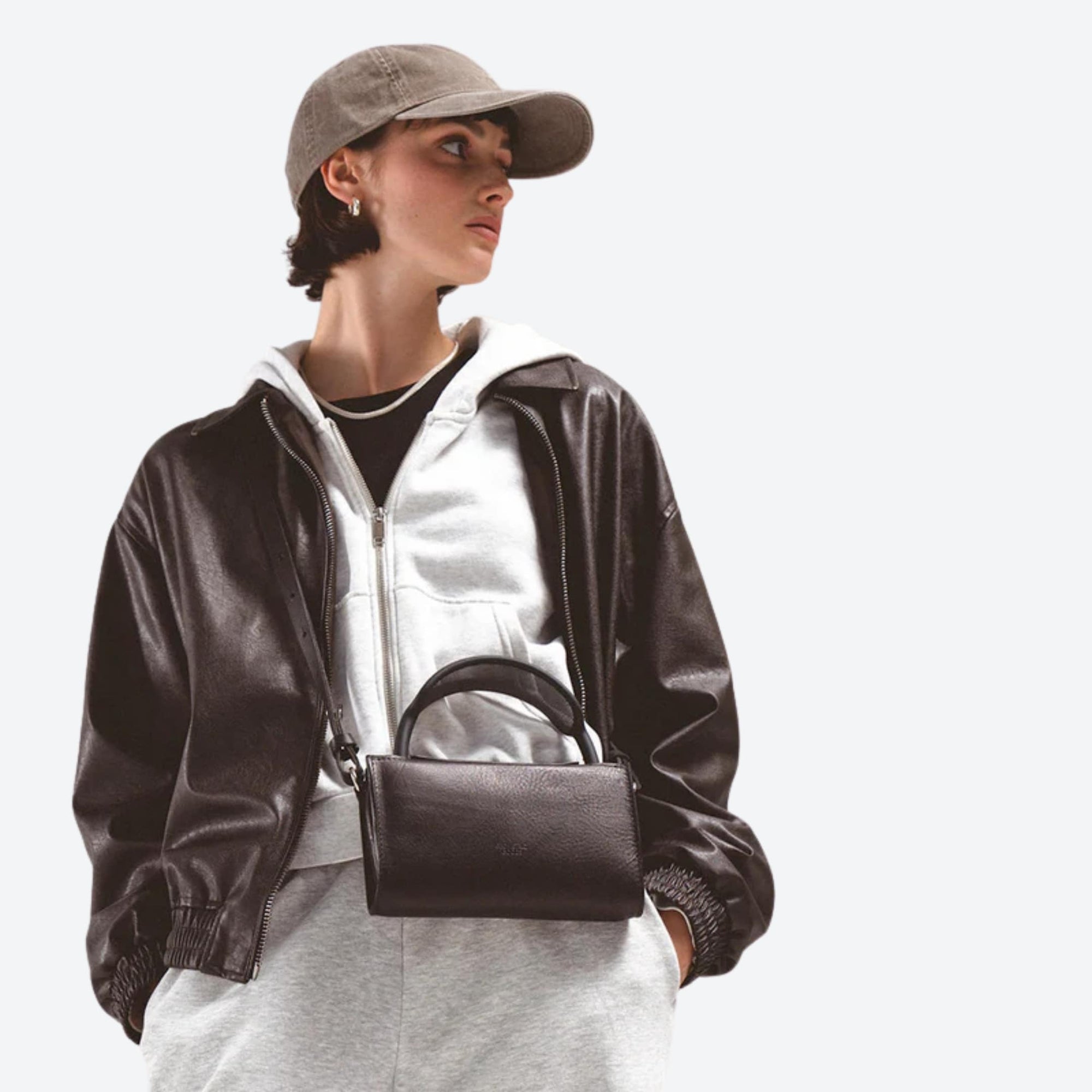 Person wearing a black leather jacket and cap, holding a black handbag on a light gray background