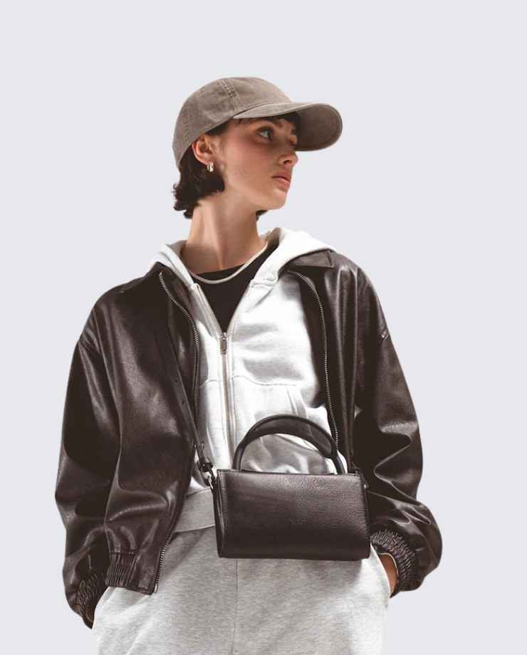 Stylish person in a brown leather jacket and cap, carrying a dark brown handbag, representing modern urban fashion and lifestyle.