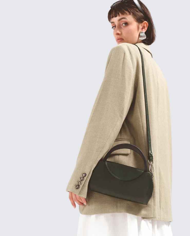 Stylish woman looking over her shoulder, wearing an oversized beige blazer and holding a dark green handbag with a unique curved handle.